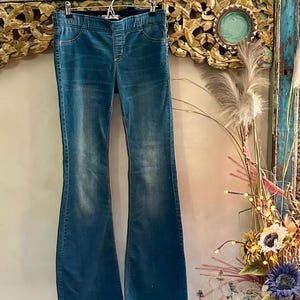 Puede incluir: Vaqueros acampanados de mezclilla azul colgados de una percha. Los vaqueros tienen una cintura alta y una abertura de pierna ancha. La mezclilla tiene un aspecto descolorido, con zonas m&aacute;s claras en los muslos y las rodillas. Los vaqueros cuelgan sobre un fondo de madera decorativo.