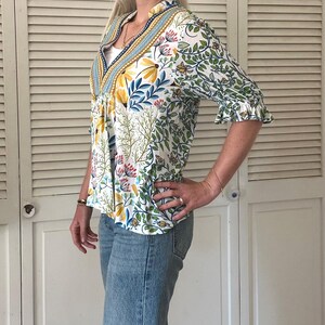 May include: A white floral print blouse with a V-neck and ruffled sleeves. The blouse features a colorful botanical pattern with yellow, blue, and pink flowers and green leaves. The blouse is paired with light blue denim jeans.