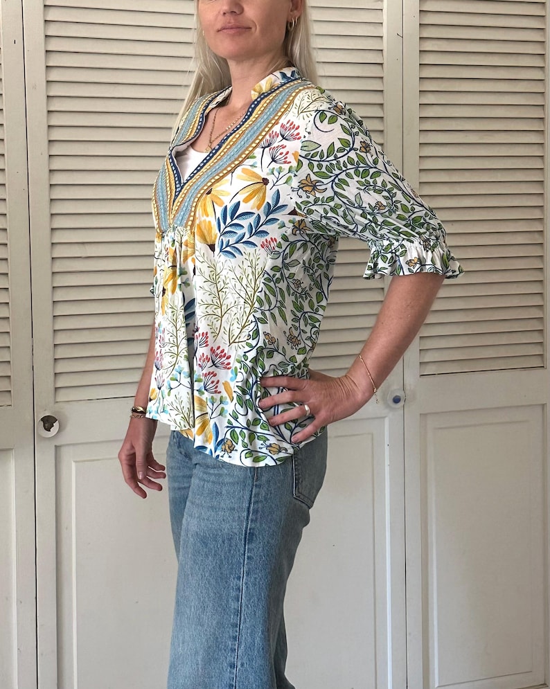 May include: A white floral print blouse with a V-neck and ruffled sleeves. The blouse features a colorful botanical pattern with yellow, blue, and pink flowers and green leaves. The blouse is paired with light blue denim jeans.