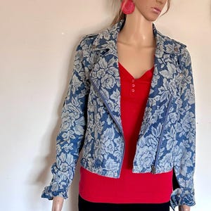 Könnte beinhalten: Eine blaue Jeansjacke mit einem weißen Blumenmuster. Die Jacke hat einen Reißverschluss und lange Ärmel. Das Model trägt ein rotes Oberteil und eine schwarze Hose. Das Design der Jacke erinnert an eine Bikerjacke.