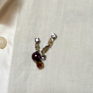 May include: A pair of decorative earrings with a cluster of beads and crystals. The earrings feature a dark purple bead, amber-colored crystals, and clear crystals. The earrings are set against a white background.