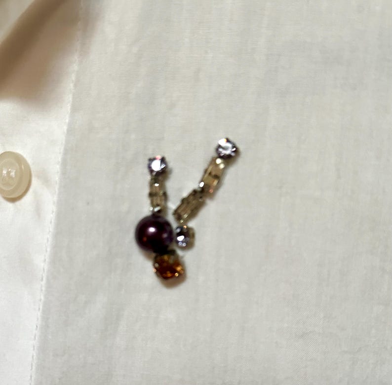 May include: A pair of decorative earrings with a cluster of beads and crystals. The earrings feature a dark purple bead, amber-colored crystals, and clear crystals. The earrings are set against a white background.