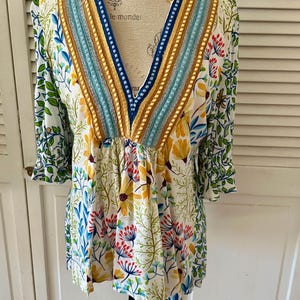 May include: A white floral print tunic top with a V-neckline. The top features a colorful floral pattern with yellow, blue, and red flowers. The neckline is embellished with blue, gold, and turquoise trim. The sleeves are three-quarter length.
