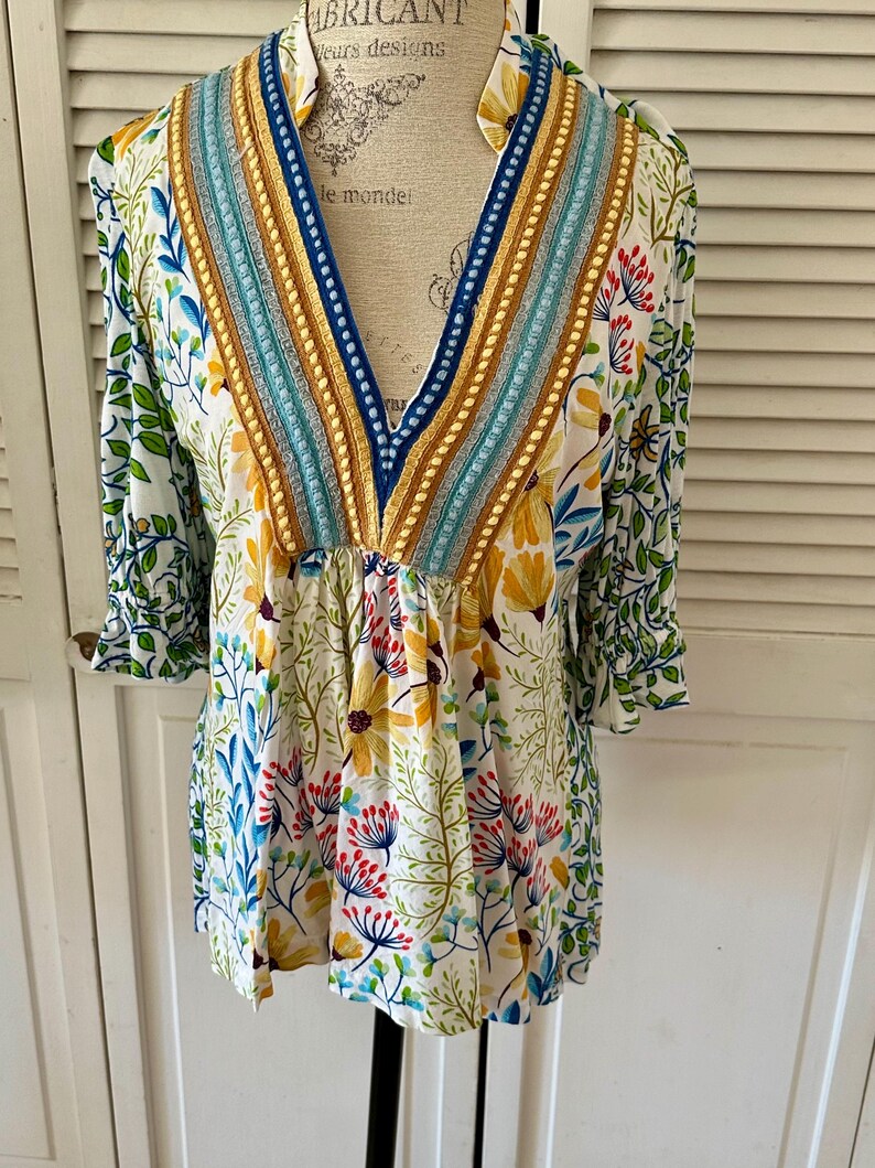 May include: A white floral print tunic top with a V-neckline. The top features a colorful floral pattern with yellow, blue, and red flowers. The neckline is embellished with blue, gold, and turquoise trim. The sleeves are three-quarter length.