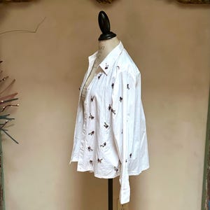 May include: White button-down shirt with long sleeves, displayed on a mannequin. The shirt is adorned with small, dark-colored embellishments scattered across the front and sleeves. The shirt has a classic collar and buttoned cuffs, suggesting a formal or semi-formal style.