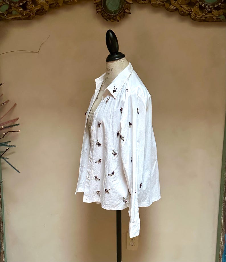 May include: White button-down shirt with long sleeves, displayed on a mannequin. The shirt is adorned with small, dark-colored embellishments scattered across the front and sleeves. The shirt has a classic collar and buttoned cuffs, suggesting a formal or semi-formal style.