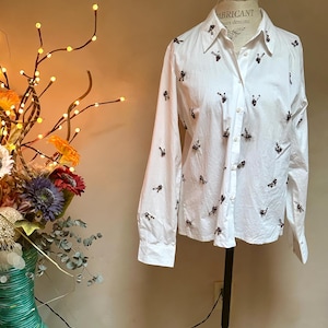 May include: A white button-down shirt with long sleeves, adorned with small, dark-colored embellishments scattered across the fabric. The shirt is displayed on a mannequin, showcasing its tailored fit and design. The collar is visible, and the shirt is buttoned up.