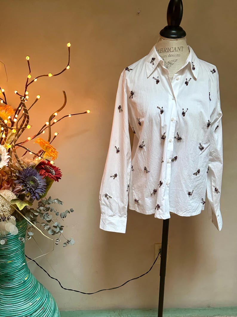 May include: A white button-down shirt with long sleeves, adorned with small, dark-colored embellishments scattered across the fabric. The shirt is displayed on a mannequin, showcasing its tailored fit and design. The collar is visible, and the shirt is buttoned up.