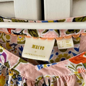 Puede incluir: Una blusa Maeve by Anthropologie con base rosa claro y un estampado floral multicolor en tonos naranja, amarillo, azul y verde. La prenda tiene una tela texturizada, un escote en V y un peque&ntilde;o borde festoneado de color naranja en el cuello.