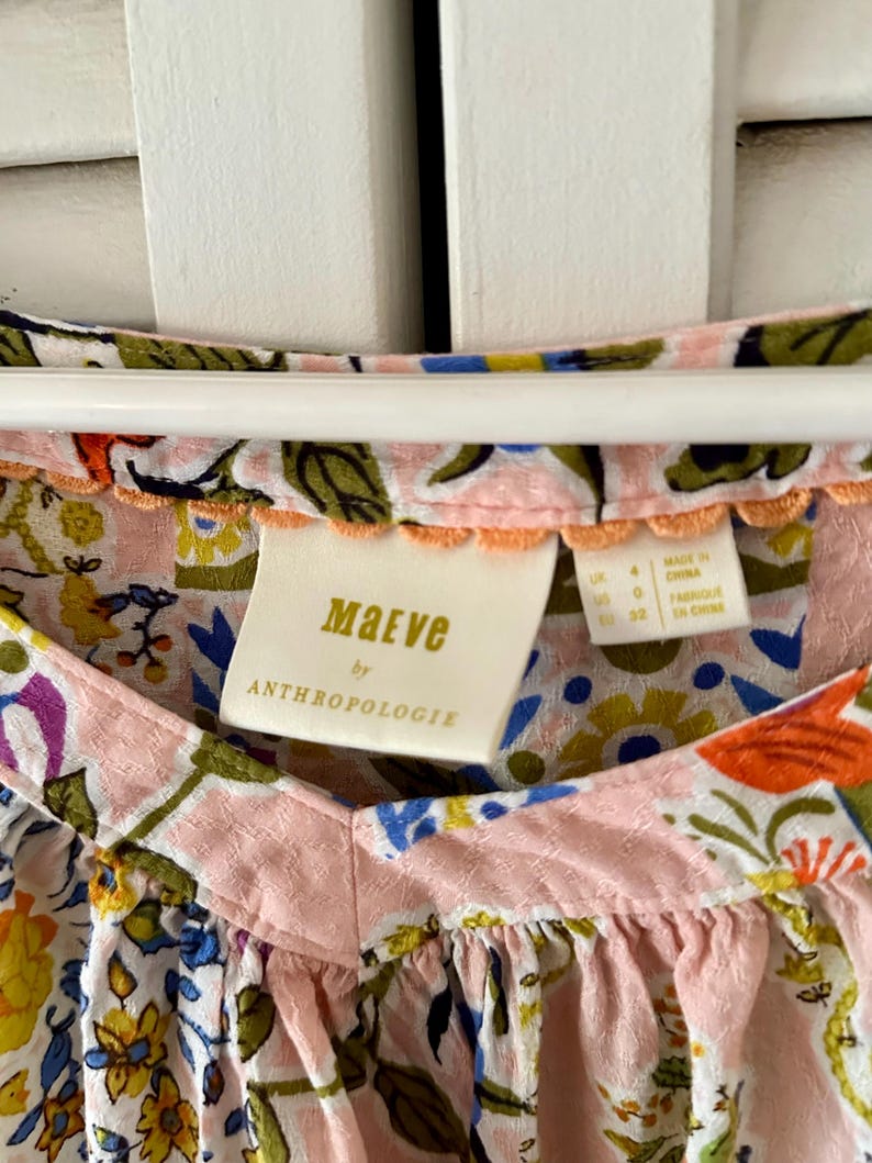 Puede incluir: Una blusa Maeve by Anthropologie con base rosa claro y un estampado floral multicolor en tonos naranja, amarillo, azul y verde. La prenda tiene una tela texturizada, un escote en V y un peque&ntilde;o borde festoneado de color naranja en el cuello.