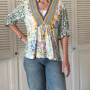 May include: A white floral print top with a V-neckline and three-quarter sleeves. The top features a blue and yellow patterned trim around the neckline and sleeves. The model is wearing blue jeans.