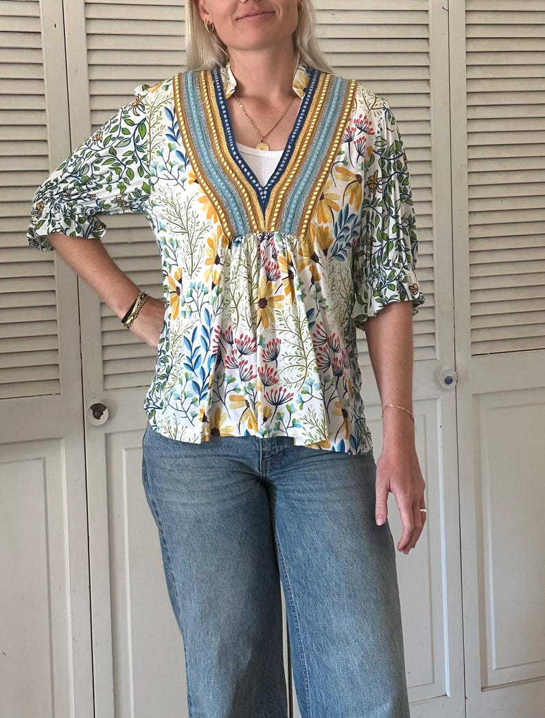 May include: A white floral print top with a V-neckline and three-quarter sleeves. The top features a blue and yellow patterned trim around the neckline and sleeves. The model is wearing blue jeans.