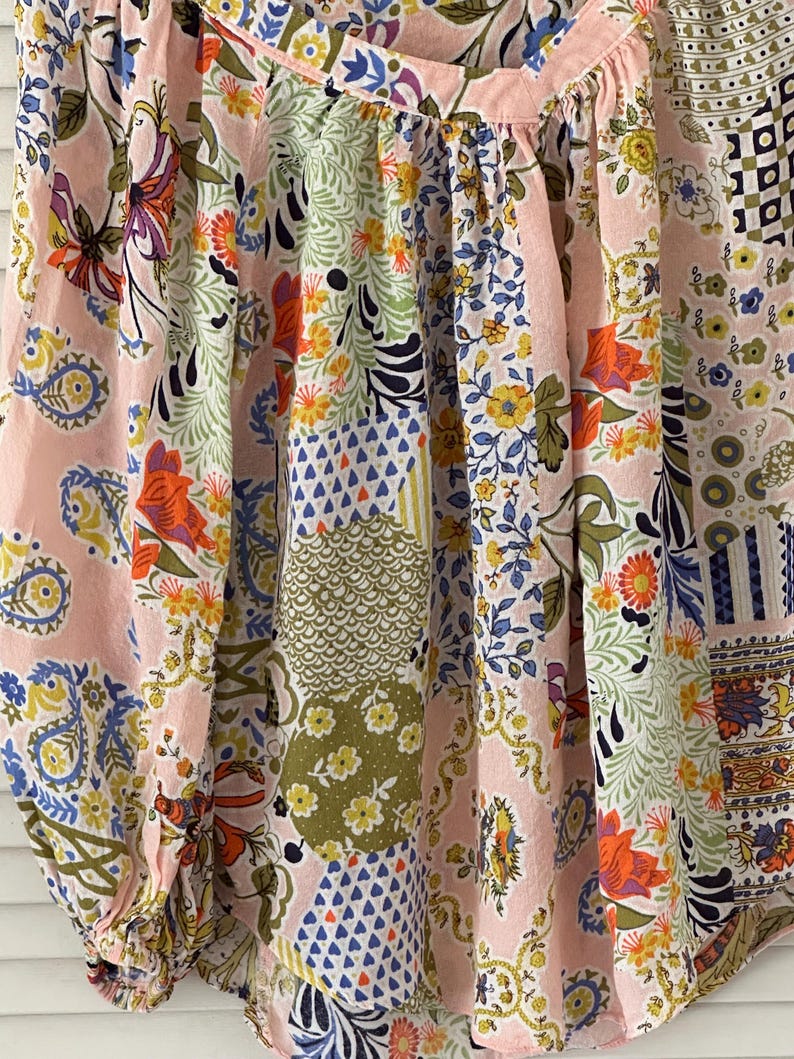 Puede incluir: Una blusa de color rosa claro con un patr&oacute;n de patchwork multicolor que presenta diversos motivos florales, de cachemira y geom&eacute;tricos en tonos naranja, amarillo, verde, azul y blanco. La prenda tiene tela fruncida en el cuello y mangas largas.
