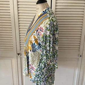 May include: A white, long-sleeved top with a colorful floral and leaf pattern. The top features a V-neckline with blue, gold, and white trim. The design includes yellow, red, and blue flowers and green leaves.