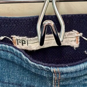 Puede incluir: Primer plano de unos pantalones cortos vaqueros azules con una cintura azul marino. Los pantalones cortos est&aacute;n colgados de una percha de metal plateado. La etiqueta dice "FP".