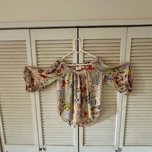 Puede incluir: Una blusa rosa claro con un patr&oacute;n de patchwork floral y geom&eacute;trico multicolor. La prenda tiene un escote en V ancho, aberturas en los hombros y mangas tres cuartos con pu&ntilde;os fruncidos, colgada en una percha de pl&aacute;stico blanca.