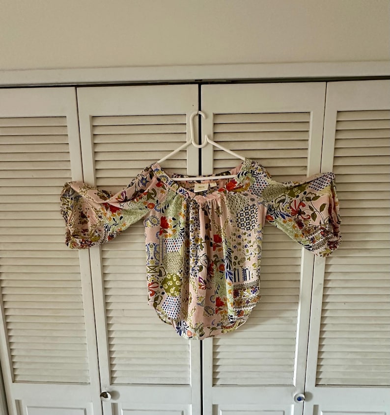 Puede incluir: Una blusa rosa claro con un patr&oacute;n de patchwork floral y geom&eacute;trico multicolor. La prenda tiene un escote en V ancho, aberturas en los hombros y mangas tres cuartos con pu&ntilde;os fruncidos, colgada en una percha de pl&aacute;stico blanca.