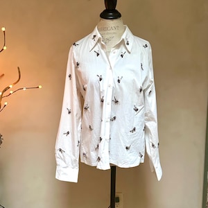 May include: White button-down shirt with long sleeves, adorned with small, dark-colored embellishments scattered across the fabric. The shirt is displayed on a mannequin, showcasing its tailored fit and design. The collar is visible, and the shirt is partially buttoned.