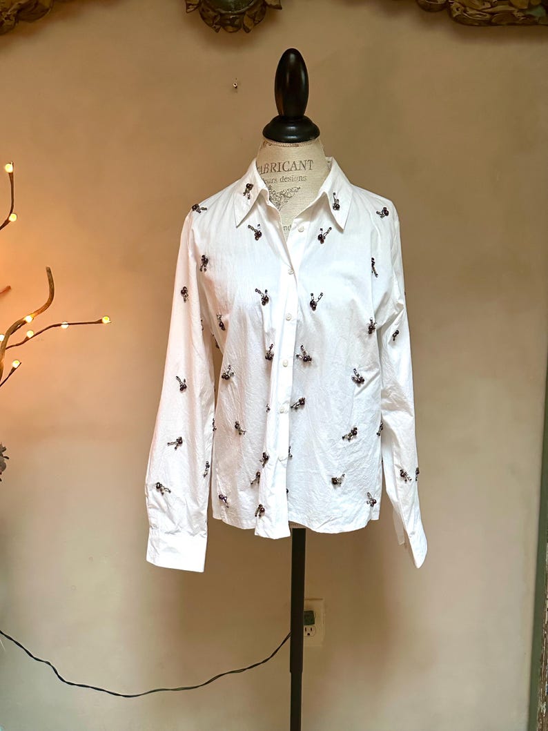 May include: White button-down shirt with long sleeves, adorned with small, dark-colored embellishments scattered across the fabric. The shirt is displayed on a mannequin, showcasing its tailored fit and design. The collar is visible, and the shirt is partially buttoned.