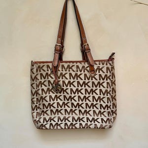 以下が含まれることがあります： ベージュのトートバッグ。茶色のアクセントとハンドルが付いています。バッグには、茶色の「MK」の文字が繰り返されるパターンがあります。ハンドルは、金色のバックルが付いた茶色の革製です。前面には小さな金属製のエンブレムがぶら下がっています。