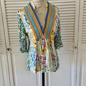 May include: A white floral print top with a V-neckline and three-quarter sleeves. The top features a colorful botanical pattern with yellow, blue, and red flowers, and green leaves. The neckline is trimmed with blue, gold, and turquoise accents.