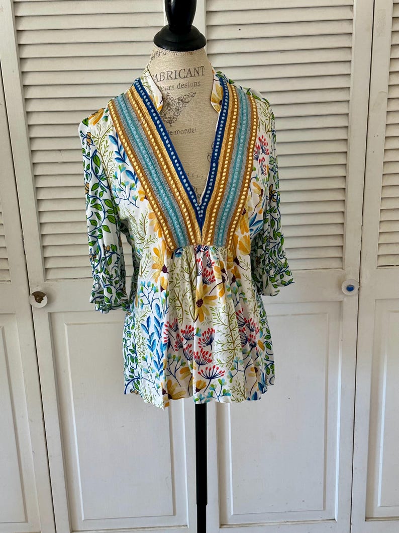 May include: A white floral print top with a V-neckline and three-quarter sleeves. The top features a colorful botanical pattern with yellow, blue, and red flowers, and green leaves. The neckline is trimmed with blue, gold, and turquoise accents.