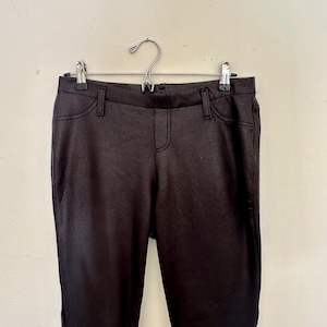 May include: Dark brown trousers with a textured, leather-look finish, suspended from a metal hanger. The trousers feature a classic five-pocket design with a button and zip fastening. They are shown against a plain, off-white wall.