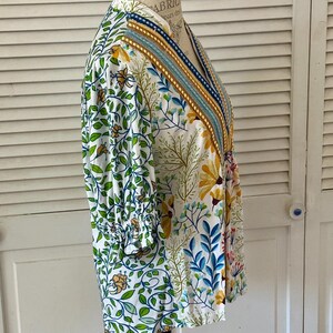 May include: A white floral print top with a V-neckline and three-quarter sleeves. The top features a colorful botanical pattern with green leaves, yellow flowers, and blue accents. The neckline is trimmed with a striped design.