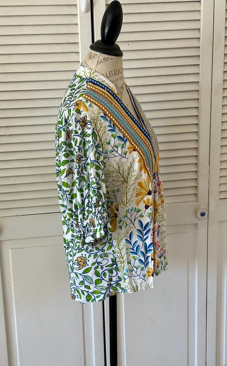 May include: A white floral print top with a V-neckline and three-quarter sleeves. The top features a colorful botanical pattern with green leaves, yellow flowers, and blue accents. The neckline is trimmed with a striped design.