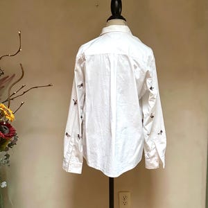 May include: White button-down shirt with long sleeves, displayed on a mannequin. The shirt features small, dark-colored embroidered details on the sleeves and upper back. The shirt is made of a lightweight fabric.