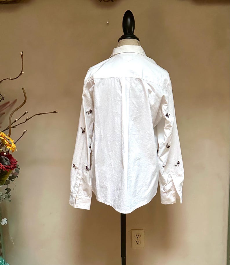 May include: White button-down shirt with long sleeves, displayed on a mannequin. The shirt features small, dark-colored embroidered details on the sleeves and upper back. The shirt is made of a lightweight fabric.