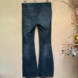 Puede incluir: Vaqueros acampanados de mezclilla azul colgados de una percha de metal. Los vaqueros tienen un dise&ntilde;o cl&aacute;sico de cinco bolsillos y una cintura alta. La mezclilla tiene un aspecto descolorido, con zonas m&aacute;s claras en los muslos y las rodillas. Los vaqueros se exhiben contra una pared de color neutro.