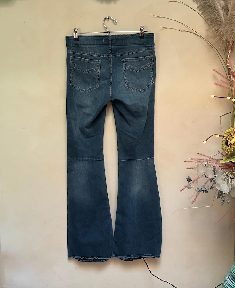 Puede incluir: Vaqueros acampanados de mezclilla azul colgados de una percha de metal. Los vaqueros tienen un dise&ntilde;o cl&aacute;sico de cinco bolsillos y una cintura alta. La mezclilla tiene un aspecto descolorido, con zonas m&aacute;s claras en los muslos y las rodillas. Los vaqueros se exhiben contra una pared de color neutro.