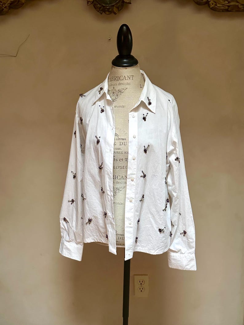 May include: White button-down shirt with long sleeves, adorned with small, dark-colored embroidered details. The shirt is displayed on a mannequin. The text "ABRICANT" and "rs designs" is visible on the mannequin.