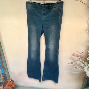 Puede incluir: Pantalones vaqueros azules con dise&ntilde;o de pernera acampanada, colgados de una percha met&aacute;lica. Los pantalones tienen un lavado oscuro con decoloraci&oacute;n m&aacute;s clara en la parte delantera de las perneras. La cintura tiene una banda el&aacute;stica oscura.