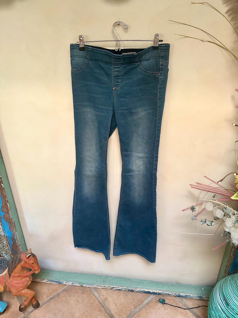 Puede incluir: Pantalones vaqueros azules con dise&ntilde;o de pernera acampanada, colgados de una percha met&aacute;lica. Los pantalones tienen un lavado oscuro con decoloraci&oacute;n m&aacute;s clara en la parte delantera de las perneras. La cintura tiene una banda el&aacute;stica oscura.