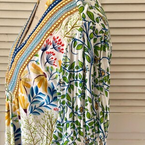 May include: White floral print top with a V-neckline. The top features a colorful embroidered trim along the neckline and a pattern of yellow flowers, blue leaves, and green vines. The sleeves are gathered at the wrist.