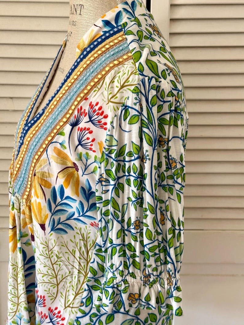 May include: White floral print top with a V-neckline. The top features a colorful embroidered trim along the neckline and a pattern of yellow flowers, blue leaves, and green vines. The sleeves are gathered at the wrist.