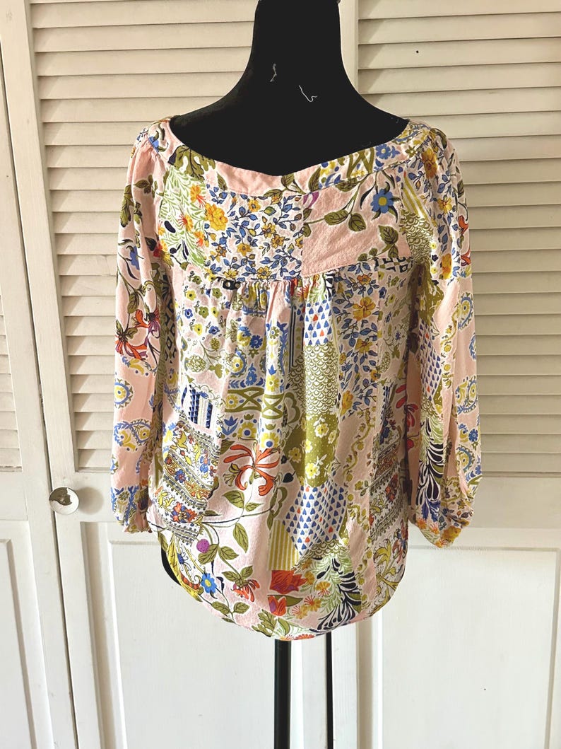 Puede incluir: Una blusa de manga larga con base rosa claro y un patr&oacute;n de patchwork multicolor con dise&ntilde;os florales, bot&aacute;nicos y geom&eacute;tricos. La prenda tiene un escote ancho y redondeado, adem&aacute;s de pu&ntilde;os fruncidos en las mangas.