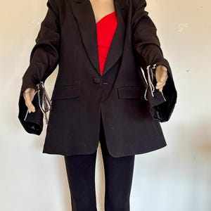 May include: Black blazer with a single-button closure, paired with black pants. The blazer features decorative silver chain accents on the sleeves. A red top is visible underneath the blazer.