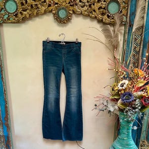 Puede incluir: Vaqueros acampanados de mezclilla azul colgados de una percha de metal. Los vaqueros tienen cintura alta y un lavado descolorido. Las perneras acampanadas crean una silueta retro. Los vaqueros se exhiben contra una pared neutra.
