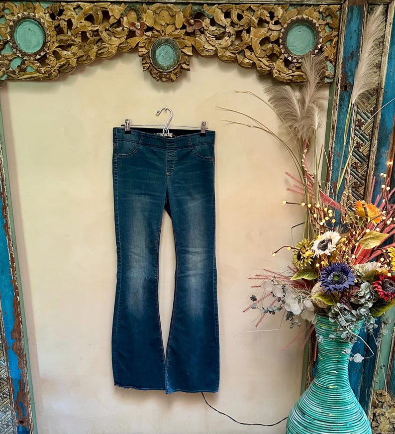 Puede incluir: Vaqueros acampanados de mezclilla azul colgados de una percha de metal. Los vaqueros tienen cintura alta y un lavado descolorido. Las perneras acampanadas crean una silueta retro. Los vaqueros se exhiben contra una pared neutra.