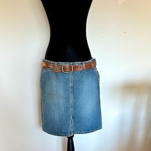 May include: A denim skirt with a brown leather belt. The belt is decorated with small, colorful embellishments. The skirt is a light blue wash and has a classic, knee-length design. The belt has a silver buckle.