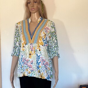 May include: A floral print blouse with a V-neckline. The blouse features a white background with yellow, blue, and red floral patterns. The neckline is accented with blue and gold trim. The sleeves are short, and the blouse is worn with black pants.