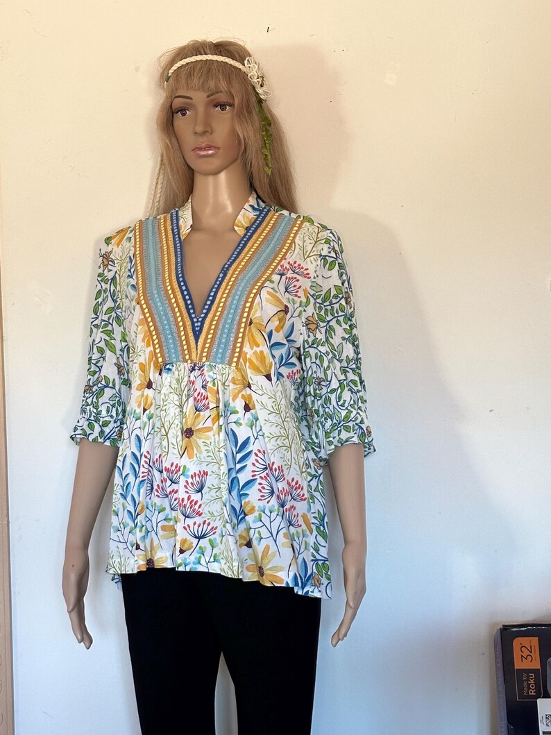 May include: A floral print blouse with a V-neckline. The blouse features a white background with yellow, blue, and red floral patterns. The neckline is accented with blue and gold trim. The sleeves are short, and the blouse is worn with black pants.