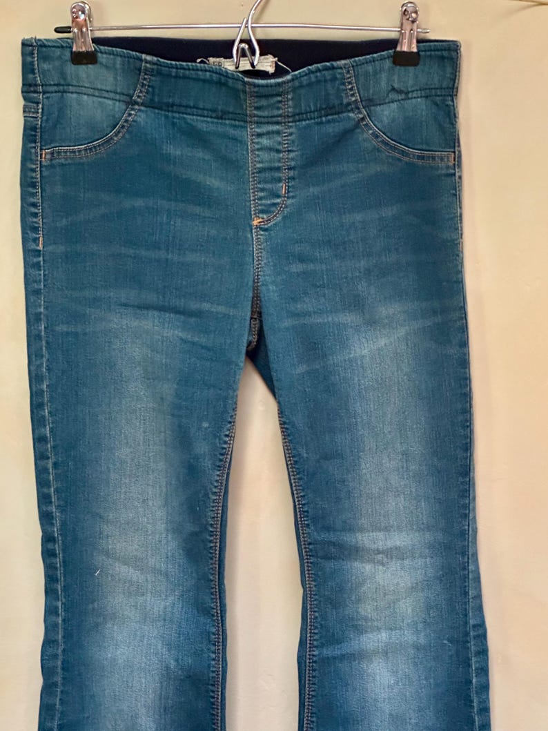 Puede incluir: Vaqueros de mezclilla azul con cintura el&aacute;stica azul oscuro y bolsillos. Los vaqueros cuelgan de una percha de metal. La mezclilla tiene un aspecto descolorido.