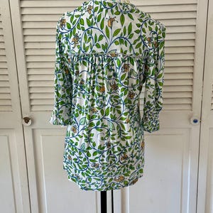 May include: White long-sleeve blouse with a botanical pattern. The design features green leaves, blue vines, and orange flowers. The blouse has a ruffled detail across the back and three-quarter sleeves.