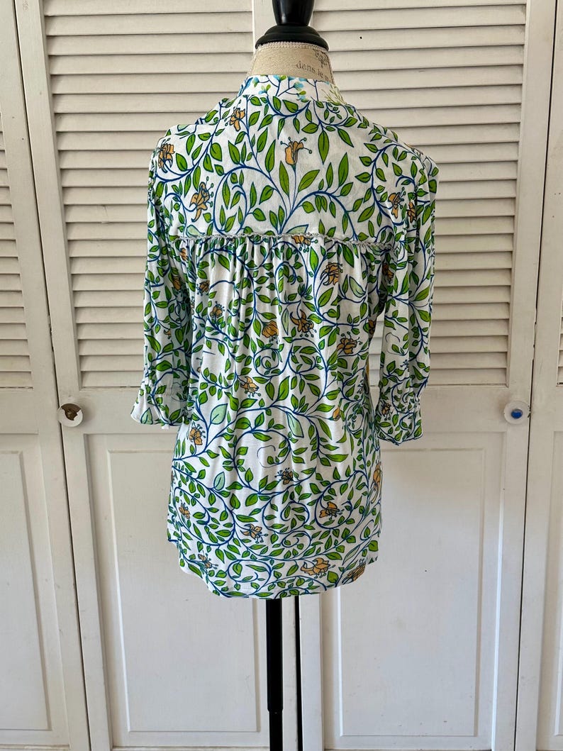 May include: White long-sleeve blouse with a botanical pattern. The design features green leaves, blue vines, and orange flowers. The blouse has a ruffled detail across the back and three-quarter sleeves.