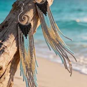 May include: Long fringe earrings featuring silver-tone metal spiral hooks and multi-strand beaded tassels. The beads are arranged in a gradient pattern of black, turquoise, and pale yellow, creating a cascading effect that hangs from the metal spirals.