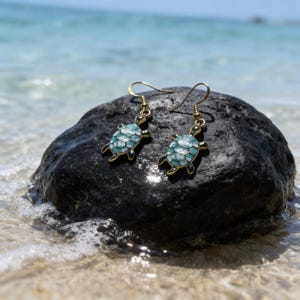 May include: A pair of gold-colored earrings featuring turquoise and white turtle designs. The earrings are displayed on a dark rock near the ocean, with the sea and sky in the background. The turtle earrings have a hook closure.